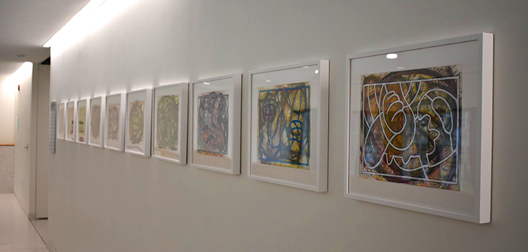 Angled photo of ten white-framed, colorful abstract paintings on a white wall.