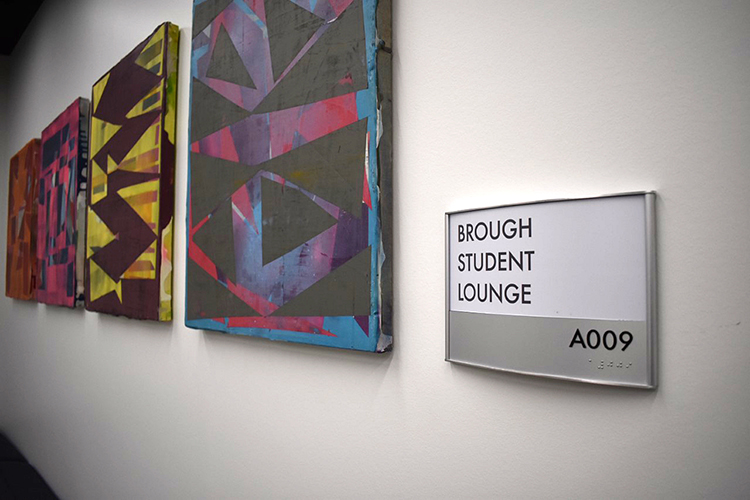  View from an angle of unframed abstract art canvasses side by side on a white wall next to a sign that reads Brough Student Lounge.