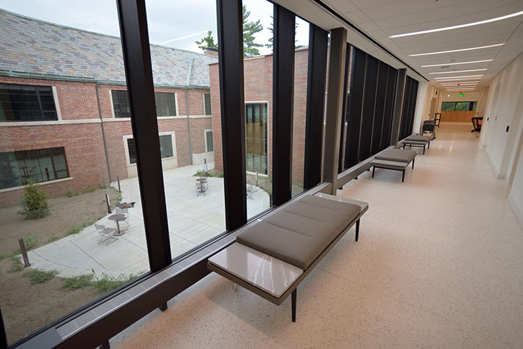 A long corridor with off-white tile floor, padded bench seating and floor-to-ceiling windows on the left, overlooking a courtyard and the original Music Building.