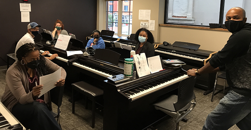 adults with masks smiling in a classroom where they are each seated at a piano