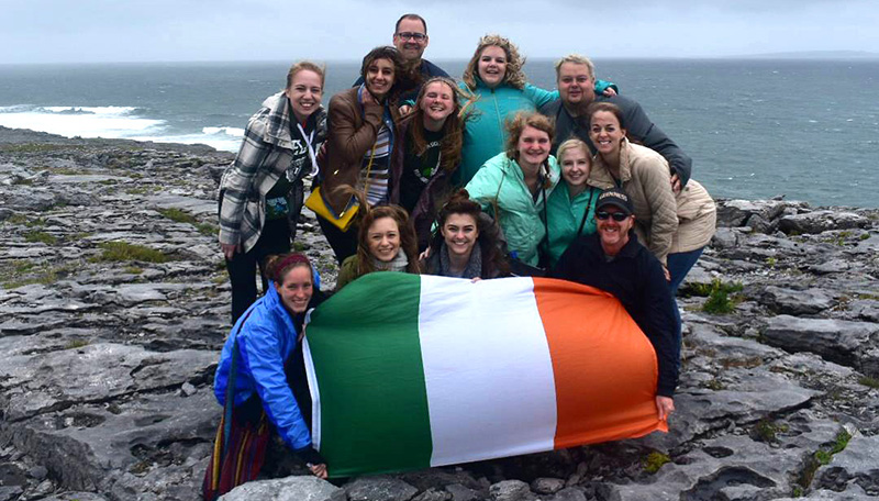 group of college students hold the flag of Ireland on a rocky shore with water in background