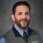 Person with dark hair and beard, wearing a blue shirt, tie and gray vest reading Michigan State.