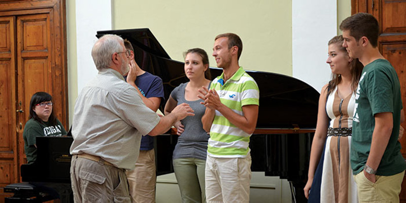 a professor speaks to a student during a lesson while three other students and a pianist look on