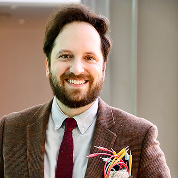 Man in brown suit and red tie smiles, has wires sticking out of the breast pocket of his jacket.