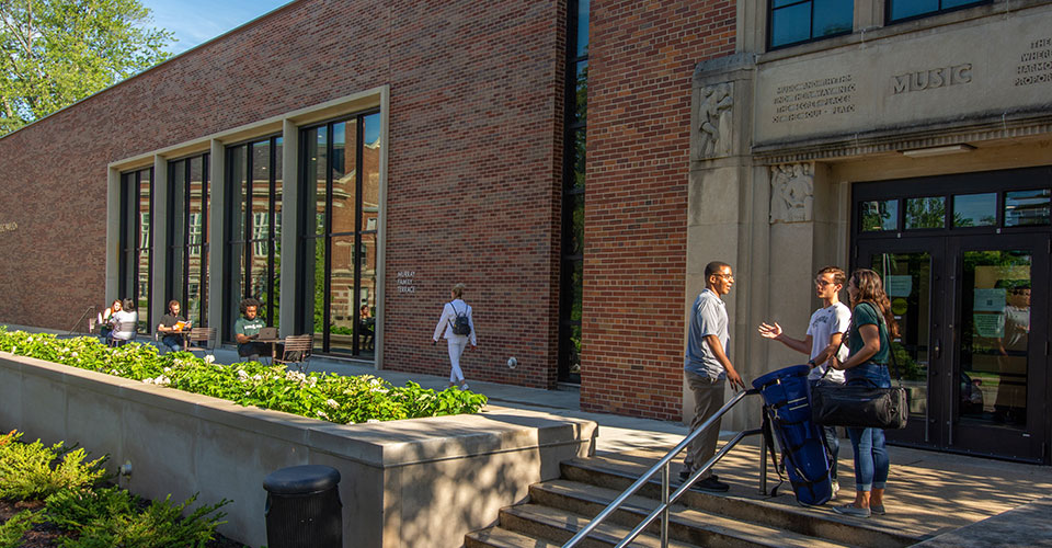 students gathered outside the front entrance of the music building, a group standing at the top of the chairs, conversing and several students seated on the terrace