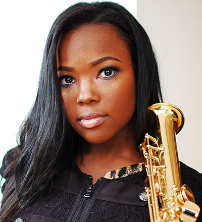 close up of woman in black outfit holding a tenor saxophone on her shoulder near her face