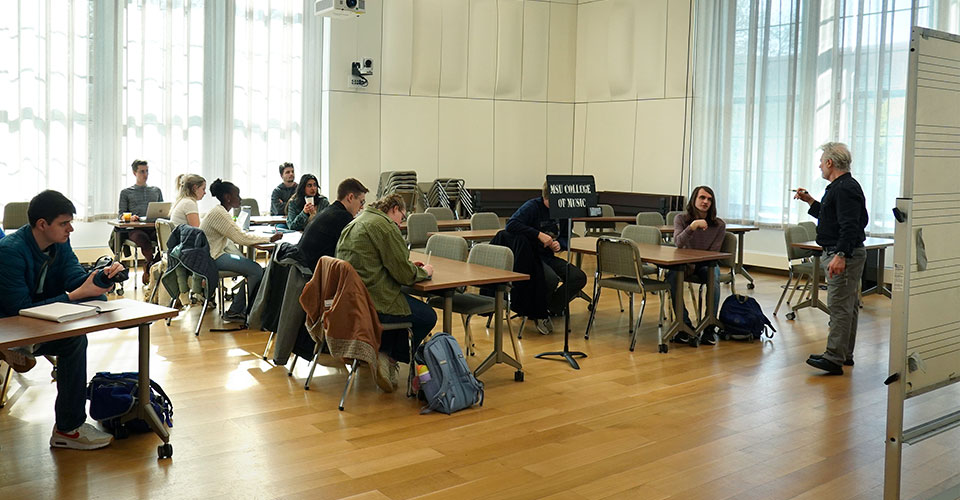 Music Theory class with about a dozen students gathered at desk-chairs listening to faculty lecture with materials referenced on board.