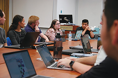 A group of students in discussion, seated around a table, each with a laptop computer in front of them.