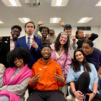 a group of 11 students, some seated, some standing, pose for a photo, smiling, laughing and making gestures.