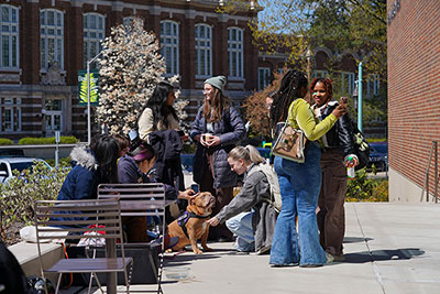 students gathered on the terrace outside of the music building. Smiling and happy students are greeting a dog.