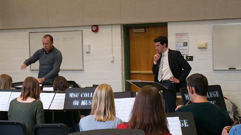 Conductor and student composer in front of wind ensemble as a new work is being rehearsed by instrumentalists