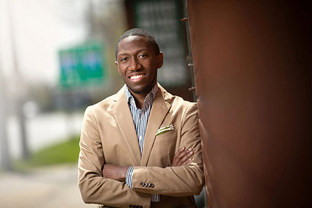 Alumnus Henry Dorn smiling and looking at camera with arms crossed, leaning up against the edge of a building with soft background of outdoors