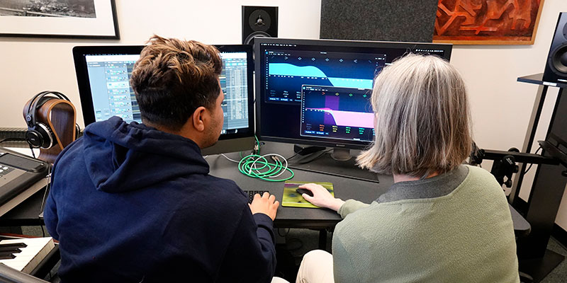 Faculty member working with a student in the composition lab, looking at software and computer screen displaying digital music