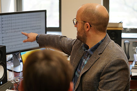 Composition faculty member references computer monitor and music with student in foreground observing