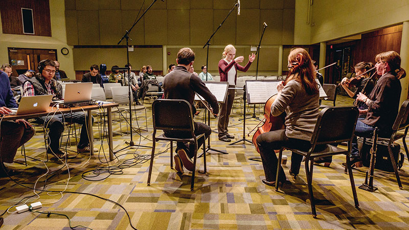 Composition student working with a guest ensemble to perform the work that she has written. Standing, conducting with instrumentalists paying attention to her cues