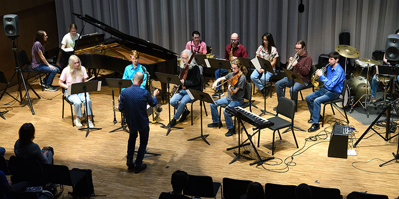 New music ensemble performing premiere of student compositions for video games. Multi instrumentalists peforming with faculty member conducting and audience members seated