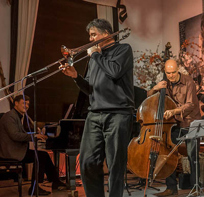 man, standing, plays trombone with two men in the background, a man on piano to the left and a man on upright bass to the right.