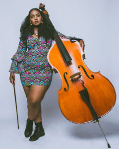 woman stands, upright bass on right leaning against her; she holds the bass bow on the left.