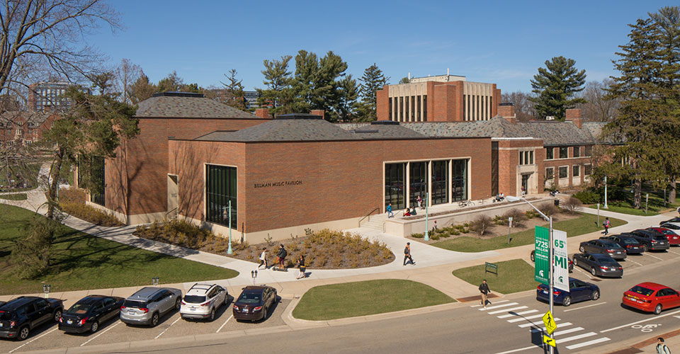 Elevated view of the entire Music Building from West Circle Drive. Sunny day with trees, gren grass, cars parked along the street and pedestrians walking in and out of building