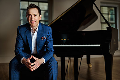 man in blue suit sits on a piano bench with grand piano in background