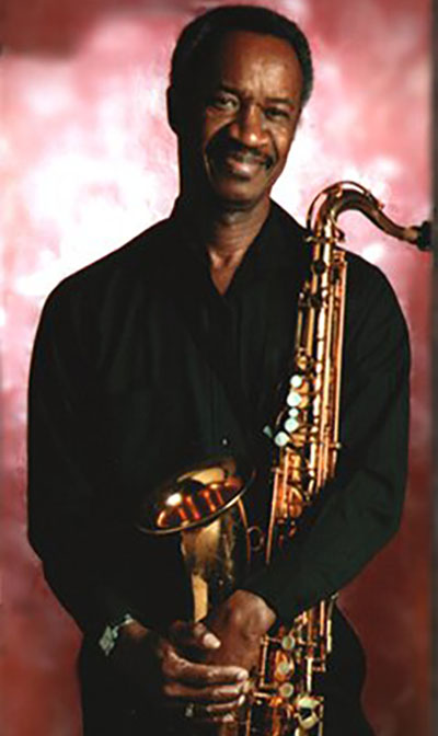 man wearing black clothing smiles and cradles a saxophone in his hands