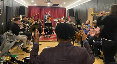 jazz band rehearsal, conductor leading with back to camera, student musicians line the walls of the room as they play