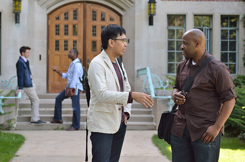 Two men speak outside on a college campus,; two other men speaking in the background on the steps of a building entrance.