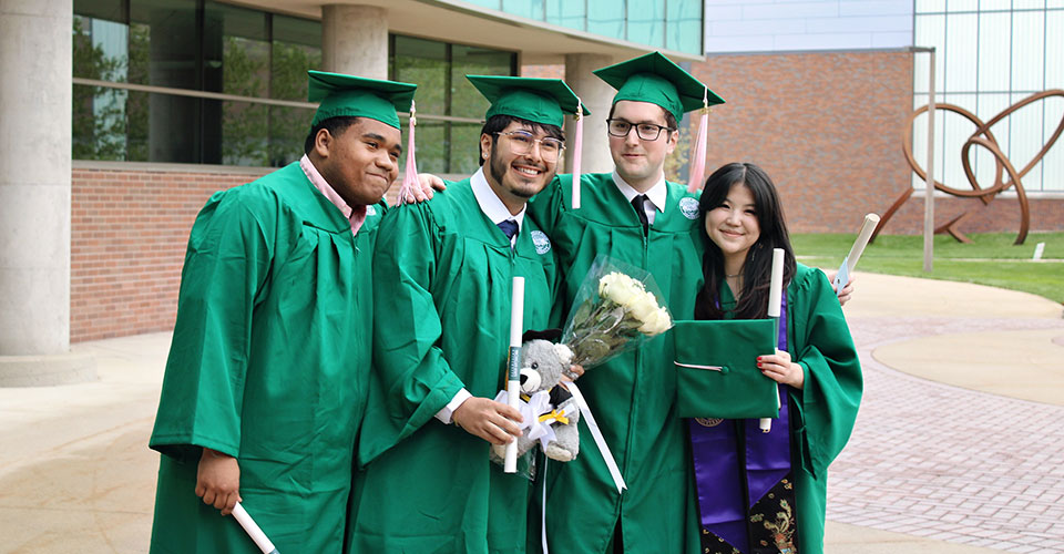 Four students gathered closely after ceremonies for a photo, wearing green cap and gown and holding diplomas