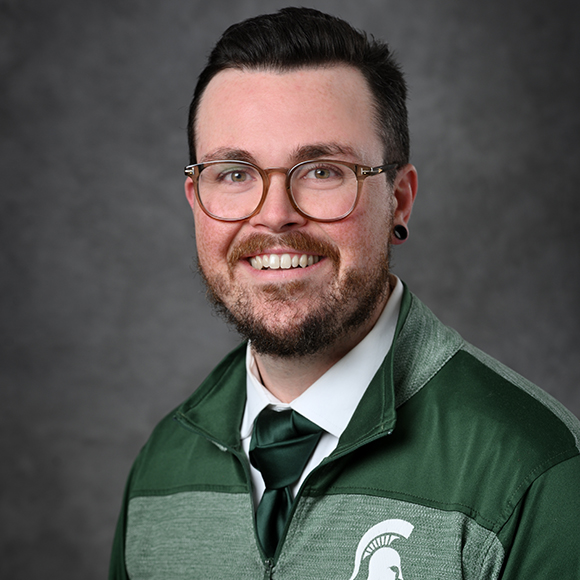 person wearing glasses, mustache and beard, white shirt with green tie, and green pullover with Michigan State University Spartan emblem.