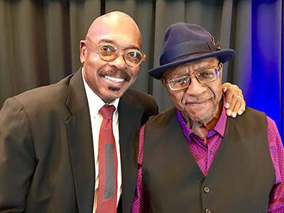 two men smile for a photo, man on left has arm around shoulder of man on right who is wearing a hat