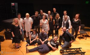 Landscape image of seventeen individuals grouped on stage for a photo. Background shows equipment, chairs and a piano.