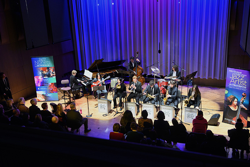 a group of nine musicians perform on jazz instrumentation with a banner on the left reading Jazz Nonets and a banner on the right reading Jazz Artist in Residence.