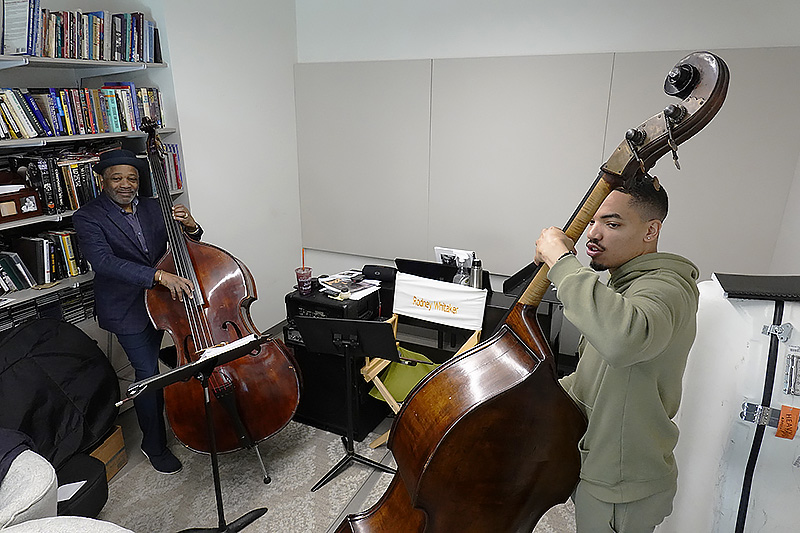 jazz professor with upright bass seated on left smiles while watching jazz student with upright bass play the instrument.