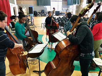 A group of upright bassists stand in a circle and rehearse together