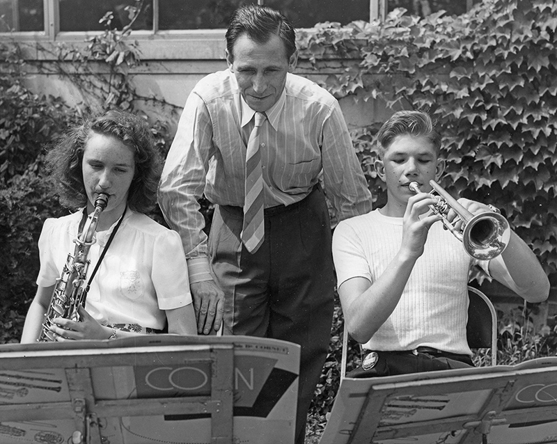 archival photo of Leonard Falcone standing in between two students playing musical instruments outdoors