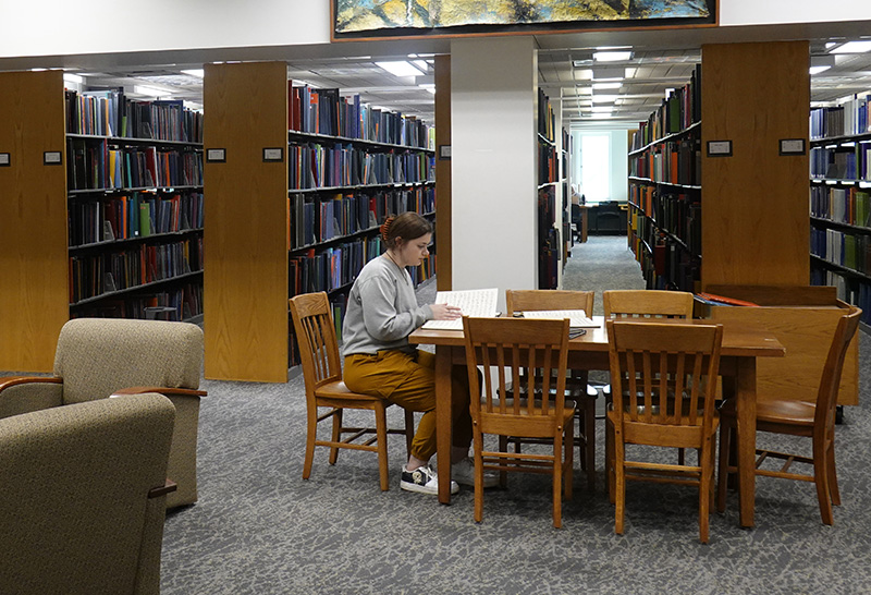 student sits in library, studying alone at a table; bookshelves in the background