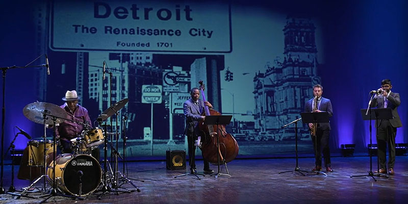 Four jazz musicians, from left, a drummer, bassist, saxophonist, and trumpeter, perform on a blue lit stage with a newspaper image in the background on a large screen that reads Detroit, The Renaissance City.