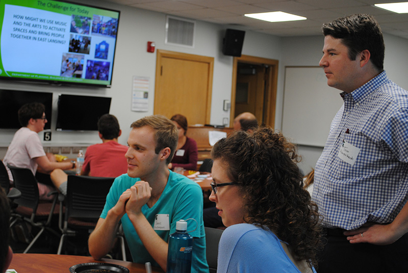 Three people, two seated, listen to someone off camera speaking, other students in background with a slid on the screen that reads challenge for today.