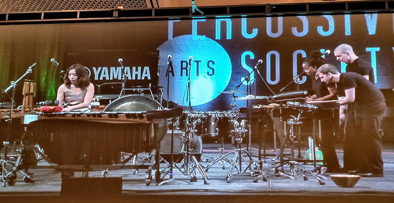 four percussionists performing on stage with a large sign in the back reading Percussion Arts Society