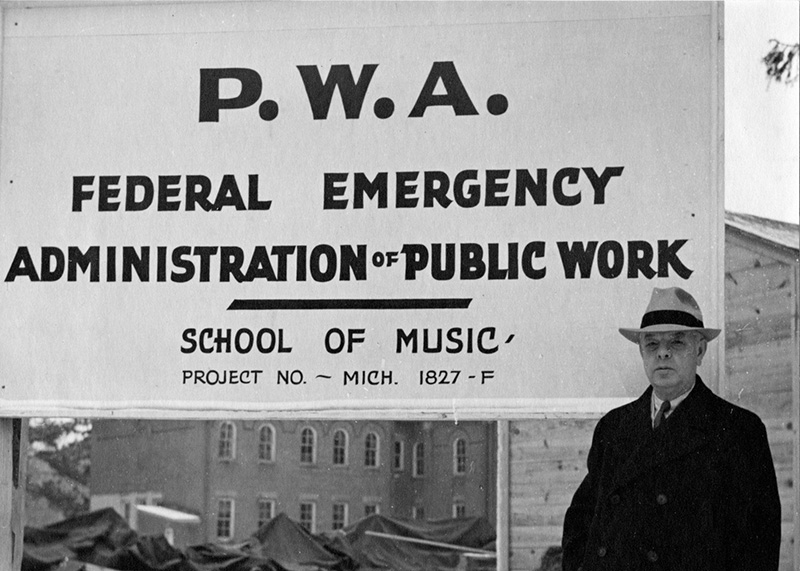 Archival photo of man standing next to a sign that reads, "PWA Federal Emergency Administration Public Work, School of Music, Project No. MICH 1827-F