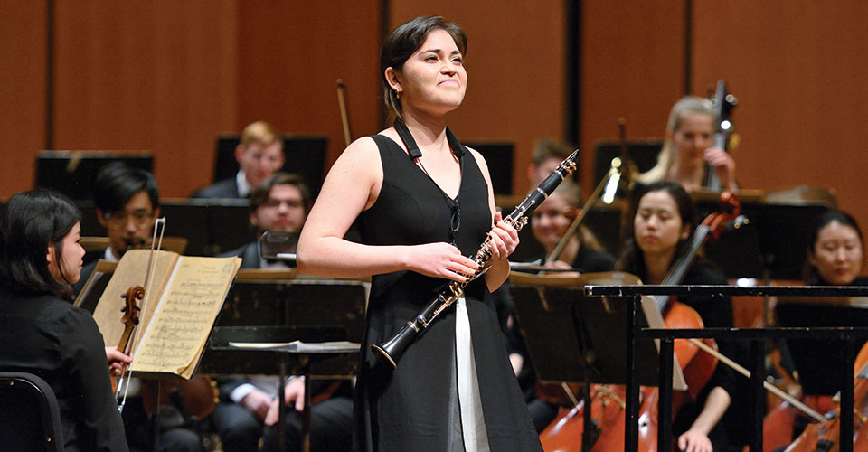 Music student holding clarinet in front of orchestra ensemble, smiling while receiving an applause from the audience.