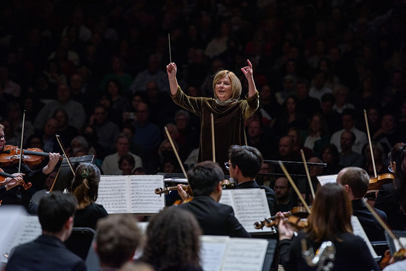 conductor motions to choir off camera, orchestra musicians in foreground