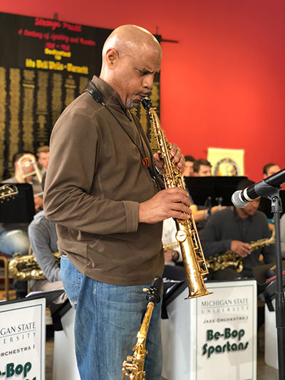 man standing and playing tenor saxophone near a microphone, other people in the background