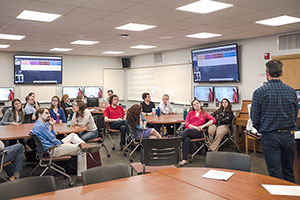 Technology classroom setting with several students listening to a lecture that has multiple display screens showing materiasl