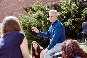 Instructor seated on a bench outside of the music building lecturing to a small group of students