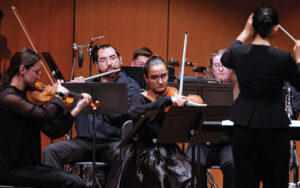 group of contemporary musicians gathered on stage, string and woodwind instrumentalists being directed by a conductor