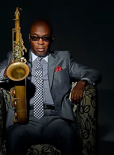 man wearing gray suit and tie with dark glasses sits and holds a saxophone