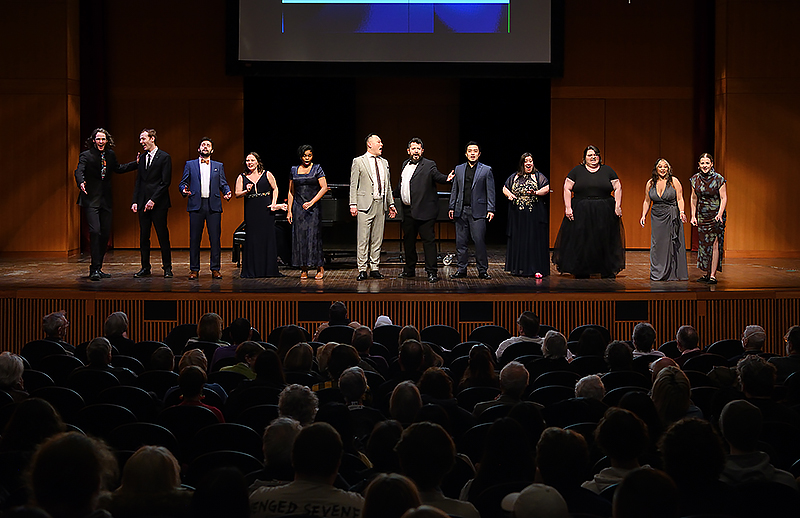 A row of 12 singers sing at the same time while performing for an audience.