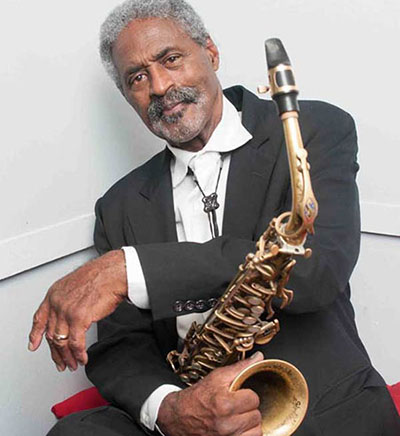 man in dark suite leans to the left and holds a saxophone with his right hand