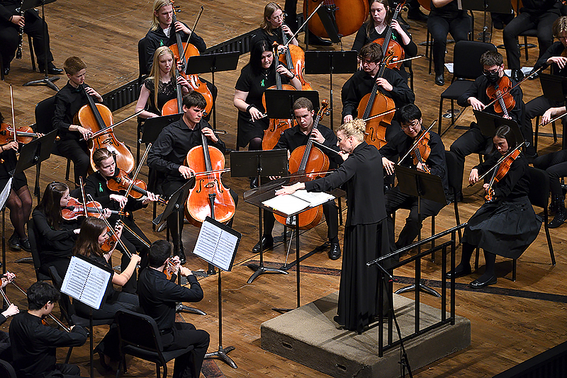 conductor on podium directs string musicians in an orchestra on stage